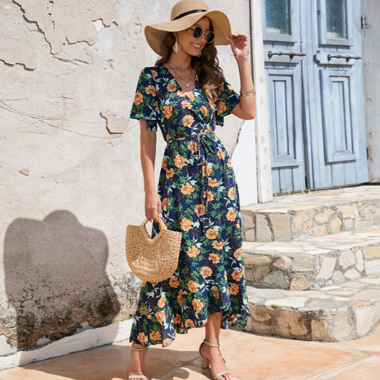 Woman in a navy blue floral summer dress with flutter sleeves, wearing a wide-brimmed straw hat and holding a woven bag, standing against a rustic Mediterranean backdrop.