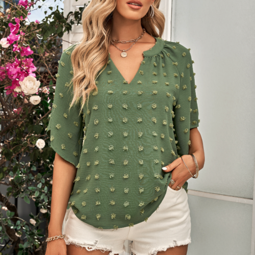 Woman wearing a green V-neck blouse with textured Swiss dot detailing and short sleeves, styled with white frayed shorts and layered jewelry in a floral outdoor setting.