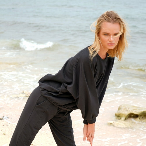 Model wearing a black oversized sweatshirt and matching joggers, leaning forward on a sandy beach with the ocean in the background and wind gently blowing her hair.