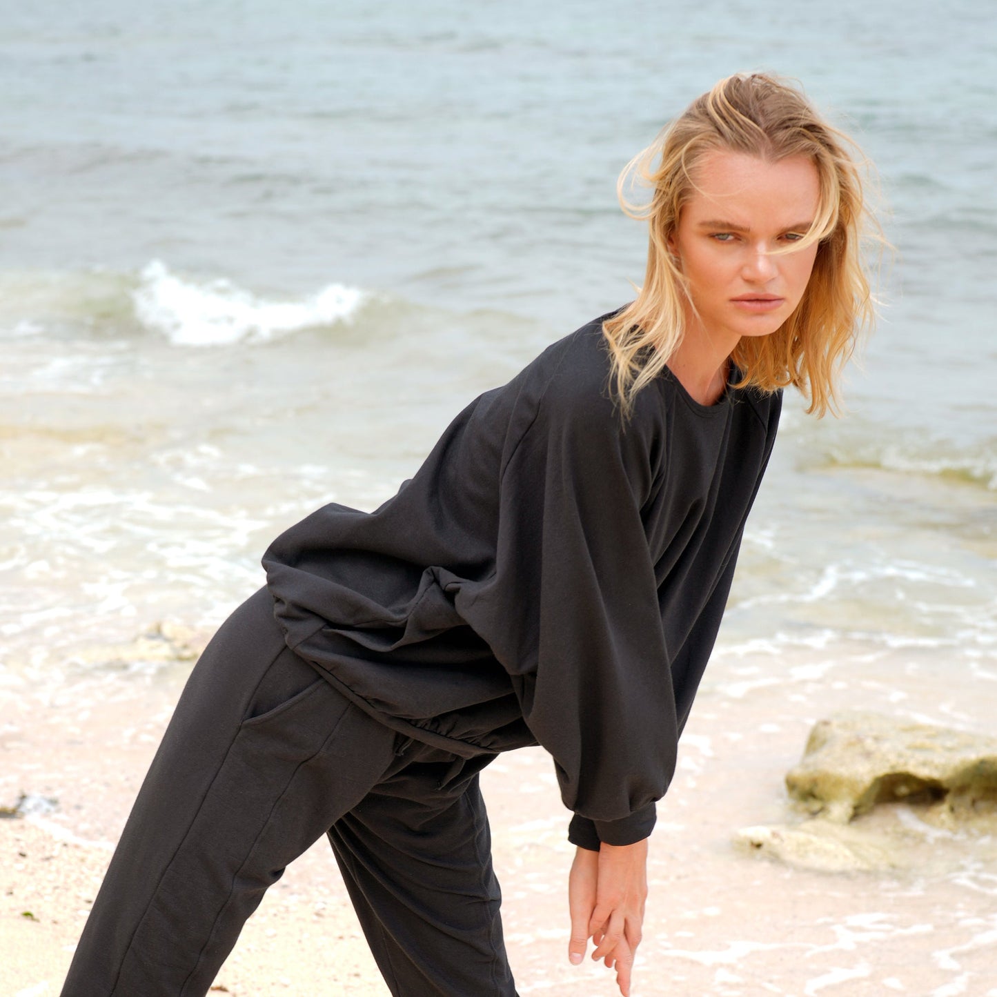 Model in loose-fitting black loungewear, leaning forward on a sandy beach with ocean waves in the background and wind blowing through her hair.