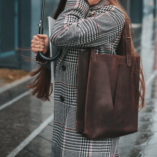 Close-up of a dark brown leather tote bag with minimalist V-cut design, carried over the shoulder of a person in a houndstooth coat, standing on a rainy city street.
