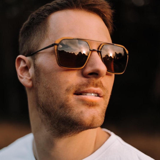 Man wearing Waidzeit handcrafted wooden sunglasses with amber-tinted lenses, reflecting a scenic sunset and forest landscape.