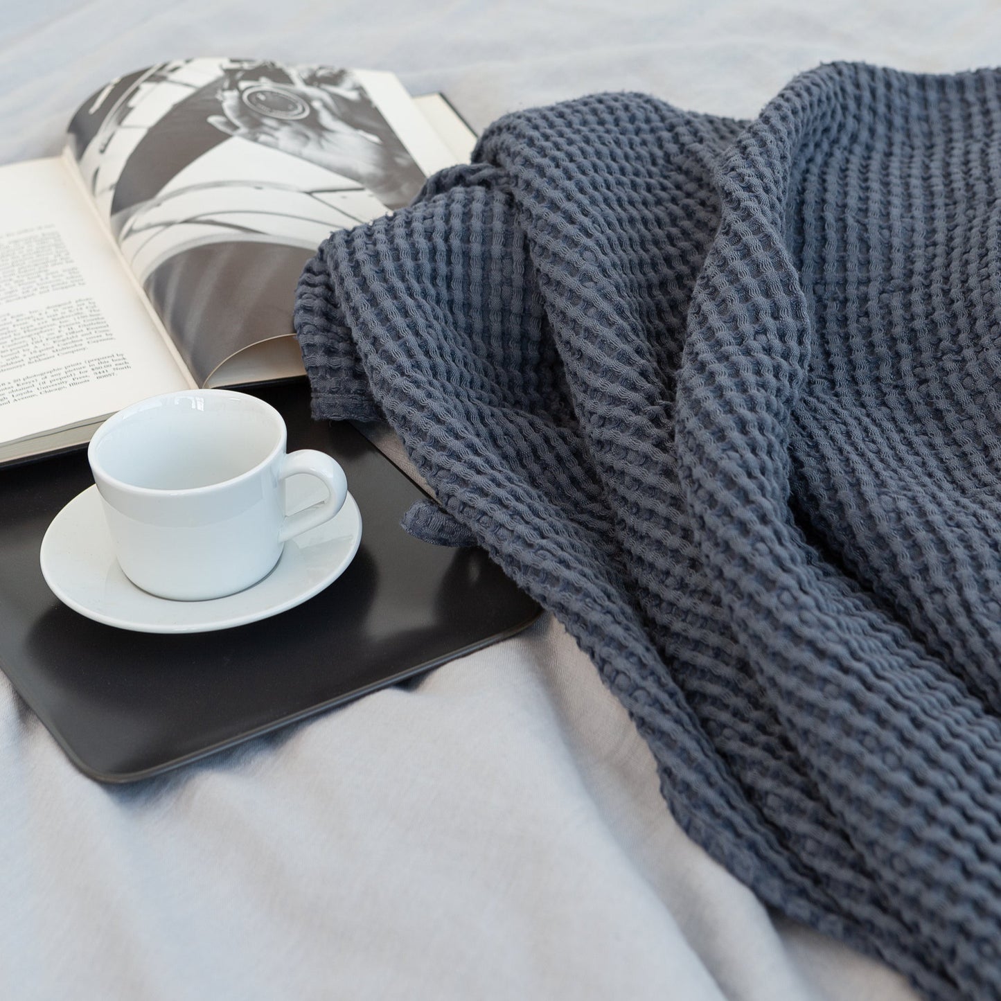 Cozy bedroom scene with dark blue waffle knit blanket, open book, and white coffee cup on a tray atop a light grey bedspread
