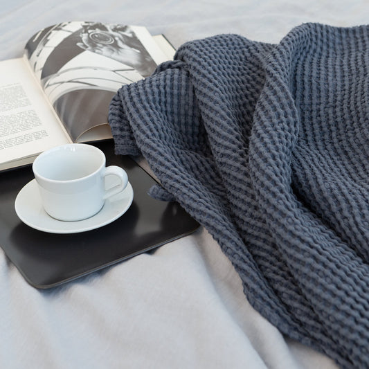 Cozy bedroom scene with dark blue waffle knit blanket, open book, and white coffee cup on a tray atop a light grey bedspread