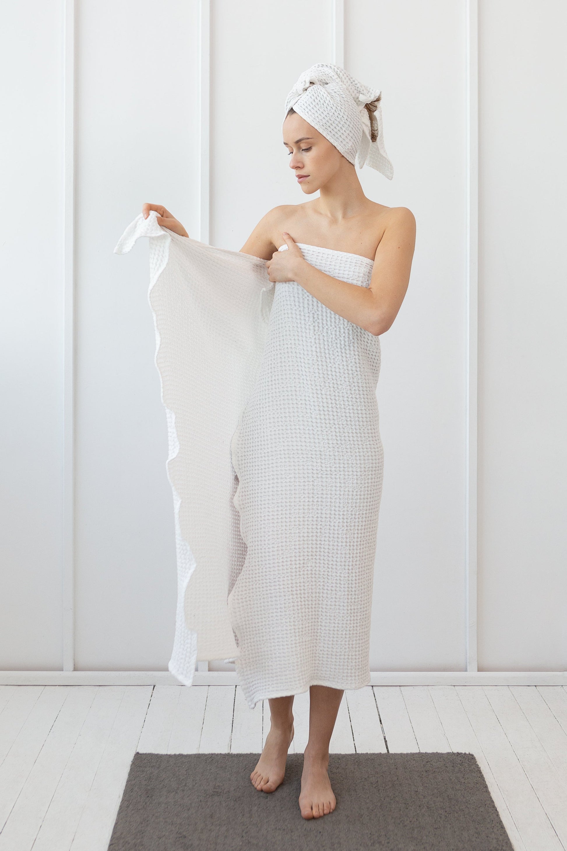 Woman in a white waffle-textured towel wrap and matching hair towel, standing in a minimal white room and holding a second towel