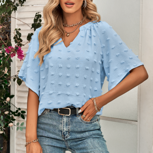 Woman wearing a light blue V-neck blouse with textured Swiss dot fabric and relaxed short sleeves, styled with blue jeans and layered necklaces in a floral outdoor setting.