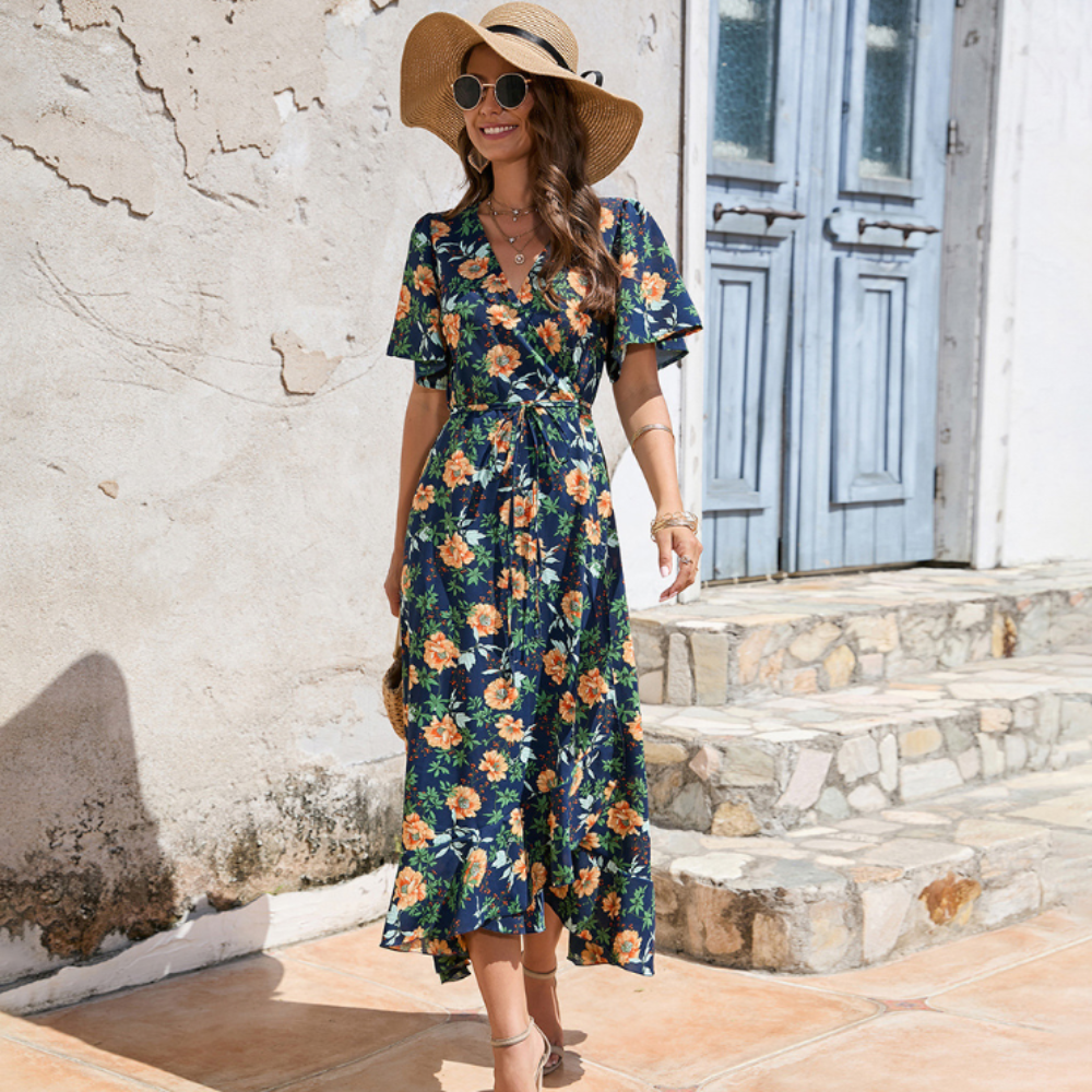 Woman wearing a navy blue floral midi dress with flutter sleeves and a tie waist, accessorized with a straw sunhat and round sunglasses, standing by a rustic stone building.
