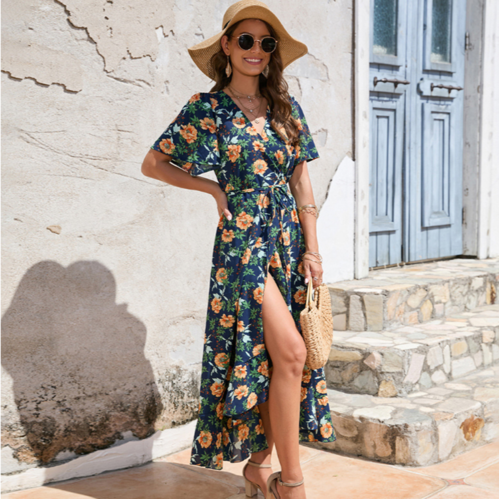 Woman in a navy floral wrap dress with front slit, wearing a straw hat, sunglasses, and holding a woven bag, posing outdoors by a sunlit stone wall.