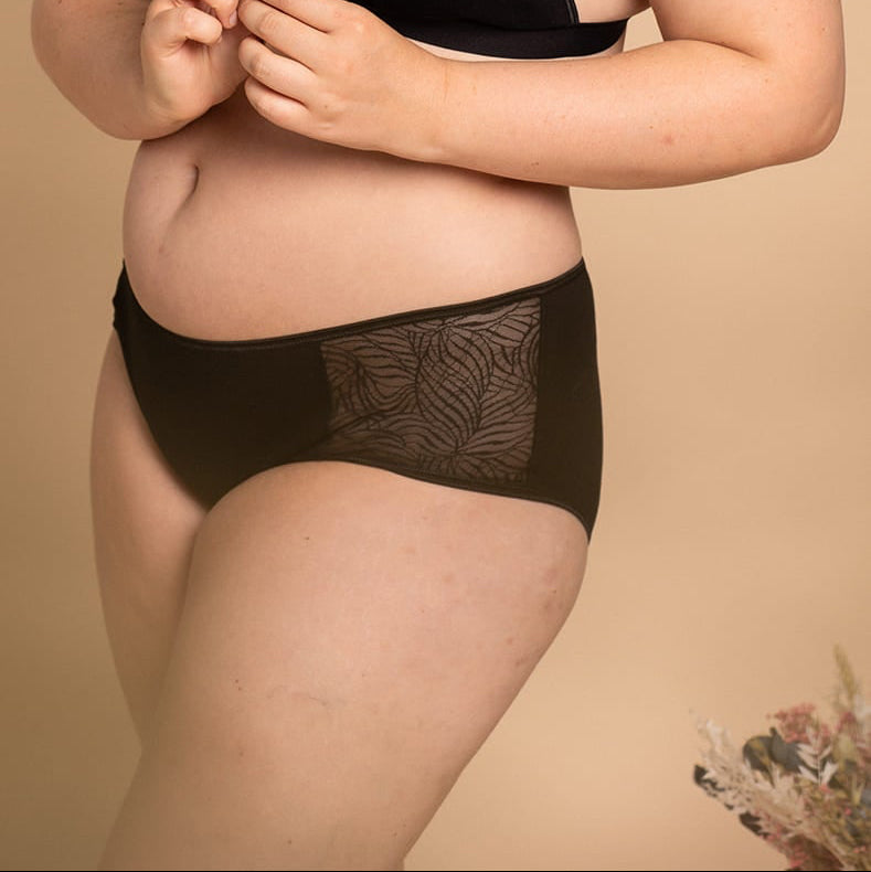 Close-up of a woman in a black lingerie set holding a small stem of blue flowers, featuring a bralette and briefs with sheer mesh panels in an abstract leaf pattern, set against a neutral beige background.
