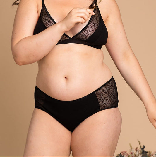 Smiling woman holding a delicate sprig of flowers, wearing a black lingerie set with soft mesh panels featuring an abstract wave pattern on the bralette and briefs, posed against a beige background.