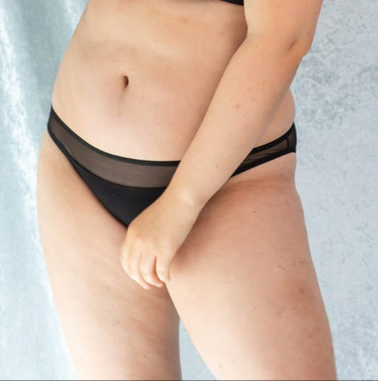 Woman posing confidently in a black sheer mesh lingerie set with a triangle bralette and bikini-style briefs, standing against a soft light blue textured backdrop.