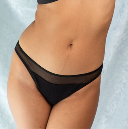 Woman stretching with arms raised overhead, wearing a sheer black lingerie set with a triangle bralette and matching bikini-style briefs, standing against a soft light blue backdrop.