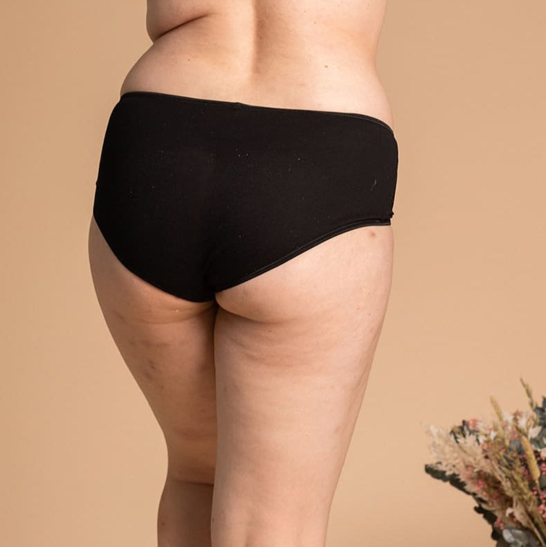 Rear view of a woman wearing black mid-rise briefs and a matching bralette with thin adjustable straps, standing against a neutral beige background with a dried floral arrangement in the corner.