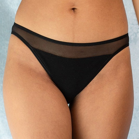Close-up front view of a woman wearing black bikini-style briefs with a sheer mesh waistband, set against a soft grey-blue background.
