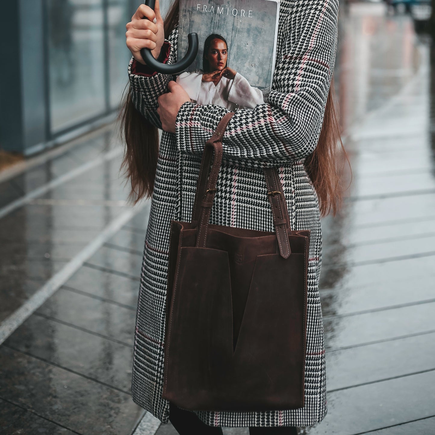Woman in a houndstooth coat holding an umbrella and a fashion magazine, wearing a premium dark brown leather tote bag with structured minimalist design, standing on a wet city pavement.