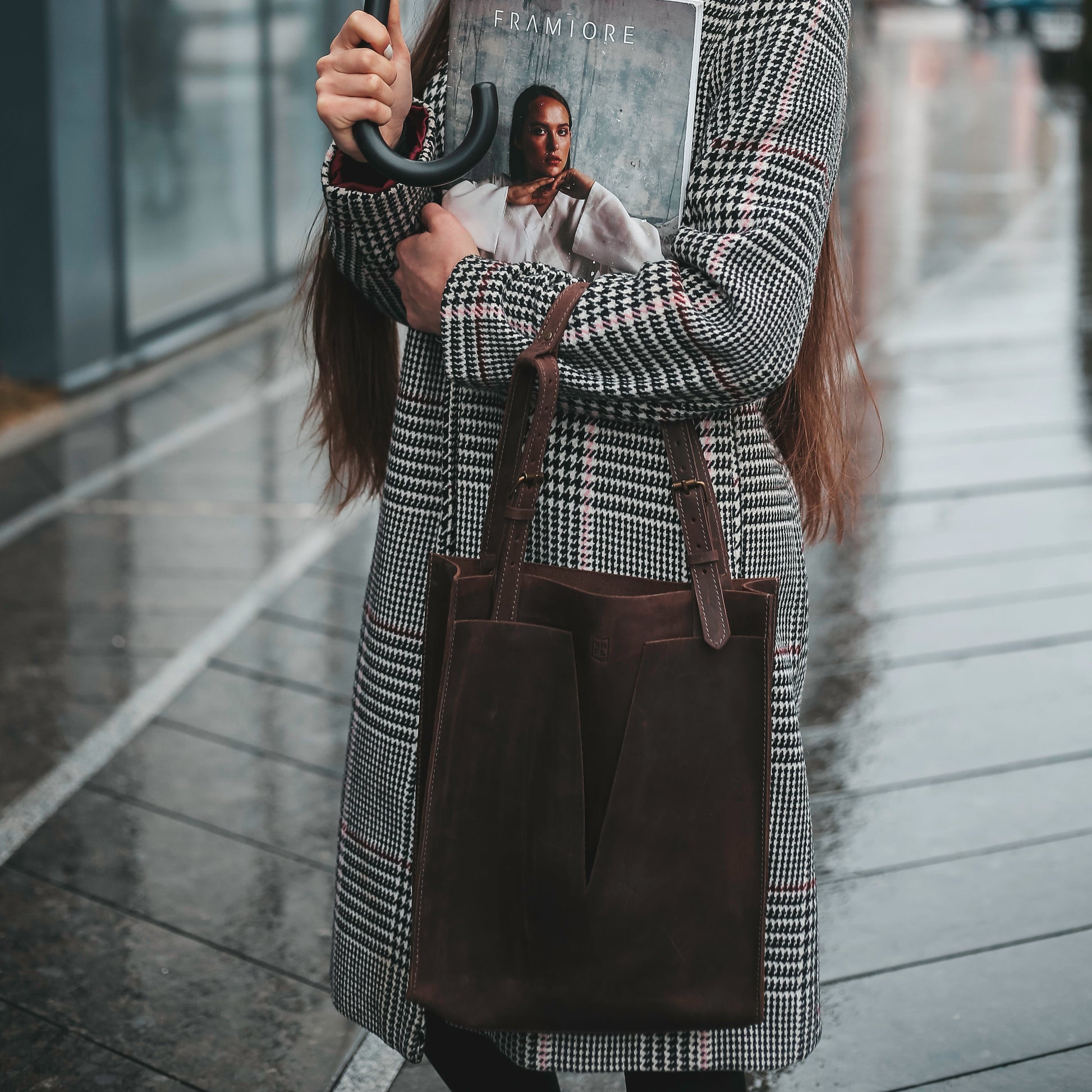 Woman in a houndstooth coat holding an umbrella and a fashion magazine, wearing a premium dark brown leather tote bag with structured minimalist design, standing on a wet city pavement.