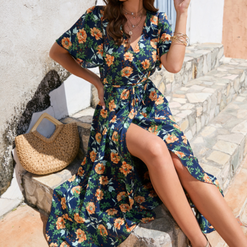 Woman seated on stone steps wearing a navy floral wrap dress with side slit, accessorized with gold jewelry and a straw handbag beside her.