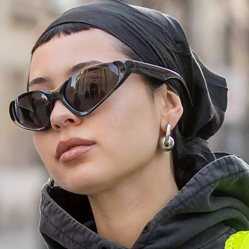 Person wearing sleek black wraparound sunglasses, a black headscarf, and a dark hoodie, accessorized with chunky silver hoop earrings in a street-style setting.