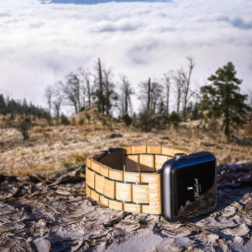 Eco-friendly Waidzeit wooden Apple Watch strap crafted from reclaimed wine barrel oak, displayed outdoors with mountain forest and cloudscape background.