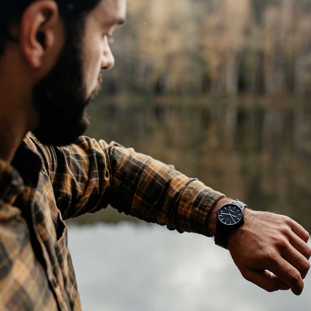 Man wearing a Waidzeit Mystic Solar watch with black dial and bog oak strap, overlooking a calm lake in an autumn forest setting.