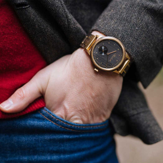 Close-up of Waidzeit men’s wristwatch with a black wooden dial and gold-toned casing, worn with a tailored jacket and jeans.