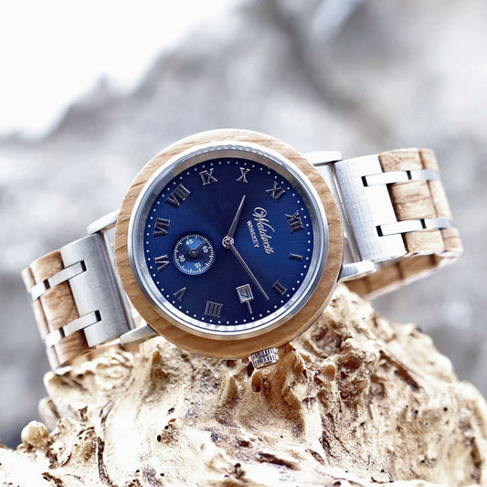 Waidzeit men's wristwatch featuring a navy blue dial, Roman numeral markers, and a wooden and stainless steel link strap, showcased on natural driftwood.