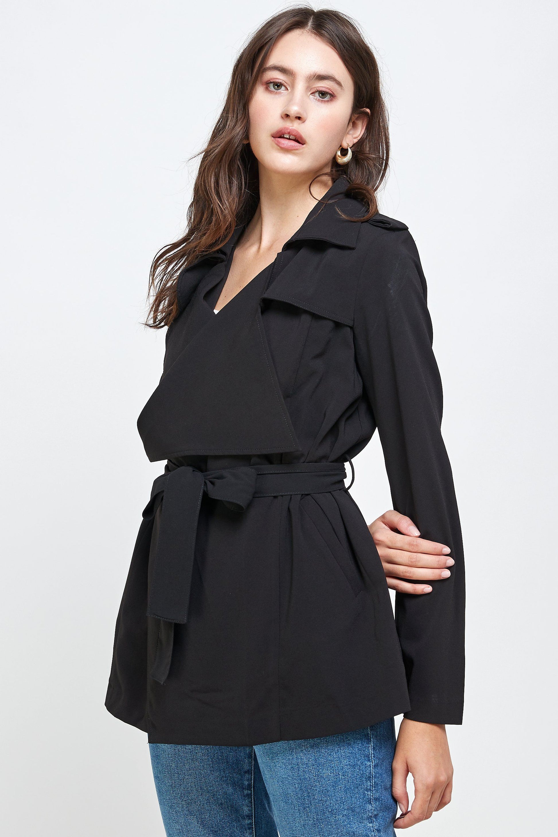 Close-up side view of a woman wearing a black belted trench jacket with lapel collar and long sleeves, styled with blue jeans for a modern casual look.
