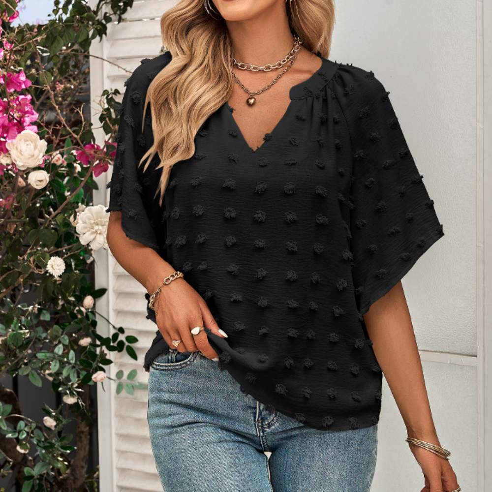 Woman in a black V-neck Swiss dot blouse with textured short sleeves, styled with denim jeans and layered necklaces, standing in front of blooming flowers.