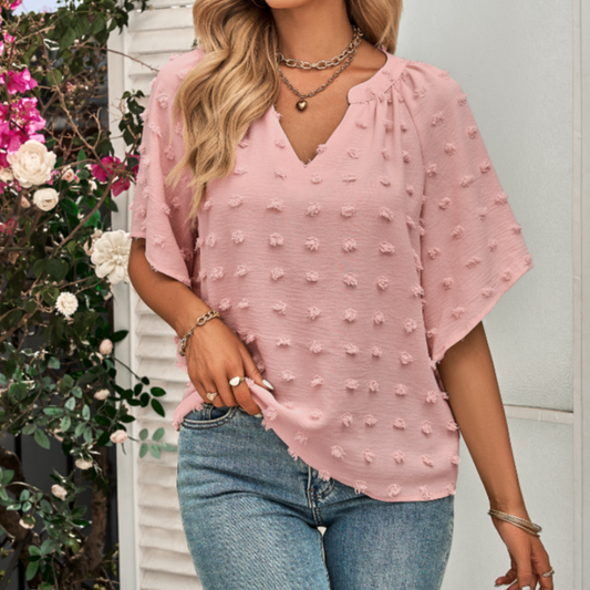 Front view of a woman wearing a pink short sleeve blouse with a V-neckline and textured Swiss dot fabric, styled casually with denim jeans and layered necklaces.