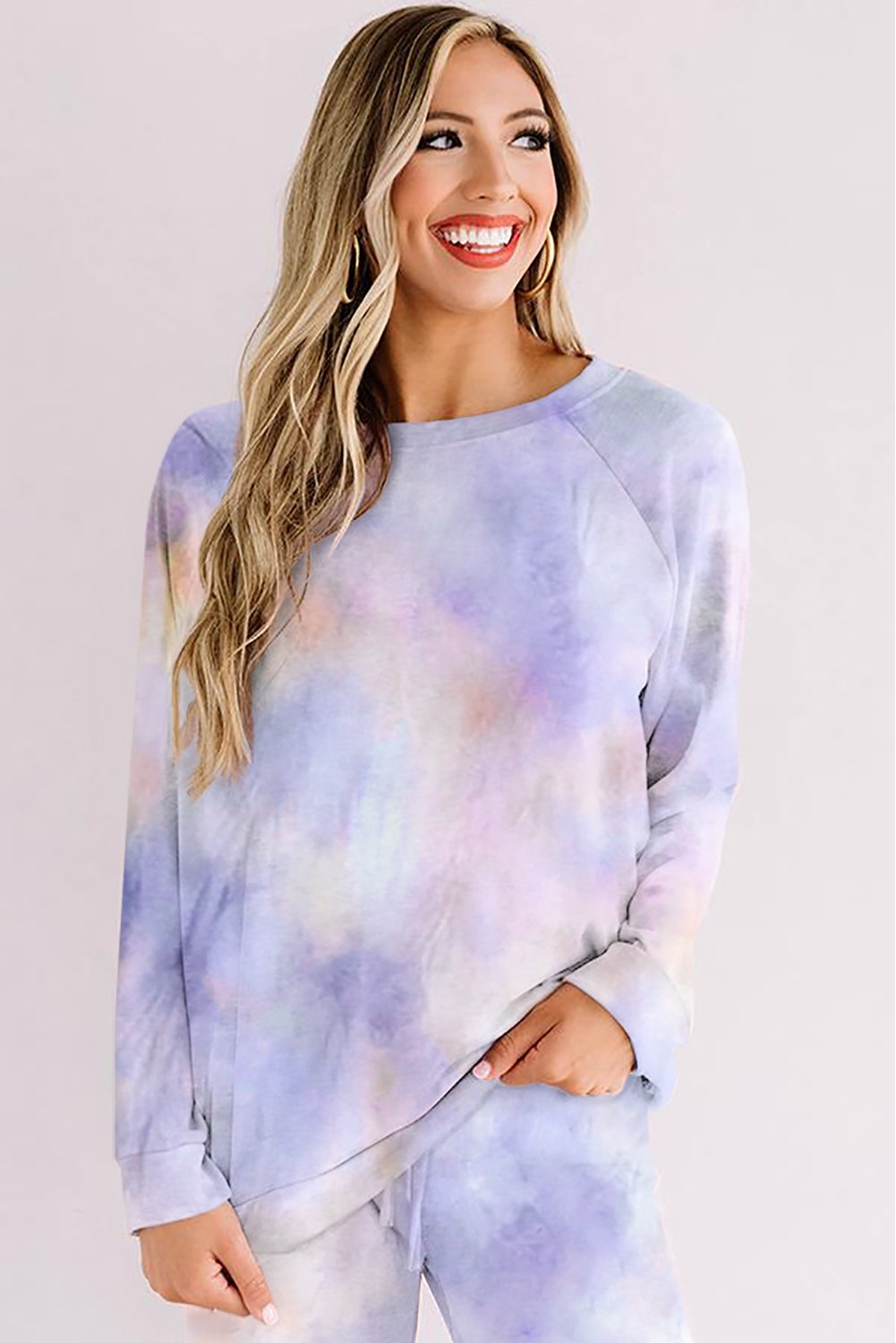 Woman smiling while wearing a pastel blue and lilac tie-dye lounge set. The outfit features a relaxed long-sleeve top and drawstring pants. She poses casually with one hand in her pocket, standing against a soft pink background.