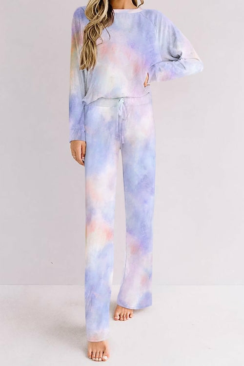 Woman wearing a pastel tie-dye loungewear set in soft shades of lavender, blue, and peach. The outfit features a long-sleeve top and drawstring wide-leg pants, styled casually as she stands barefoot against a plain light background.