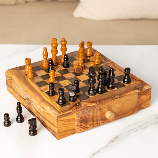 Handcrafted wooden chess set with natural and black pieces on a square board featuring integrated storage drawers, placed on a marble surface.