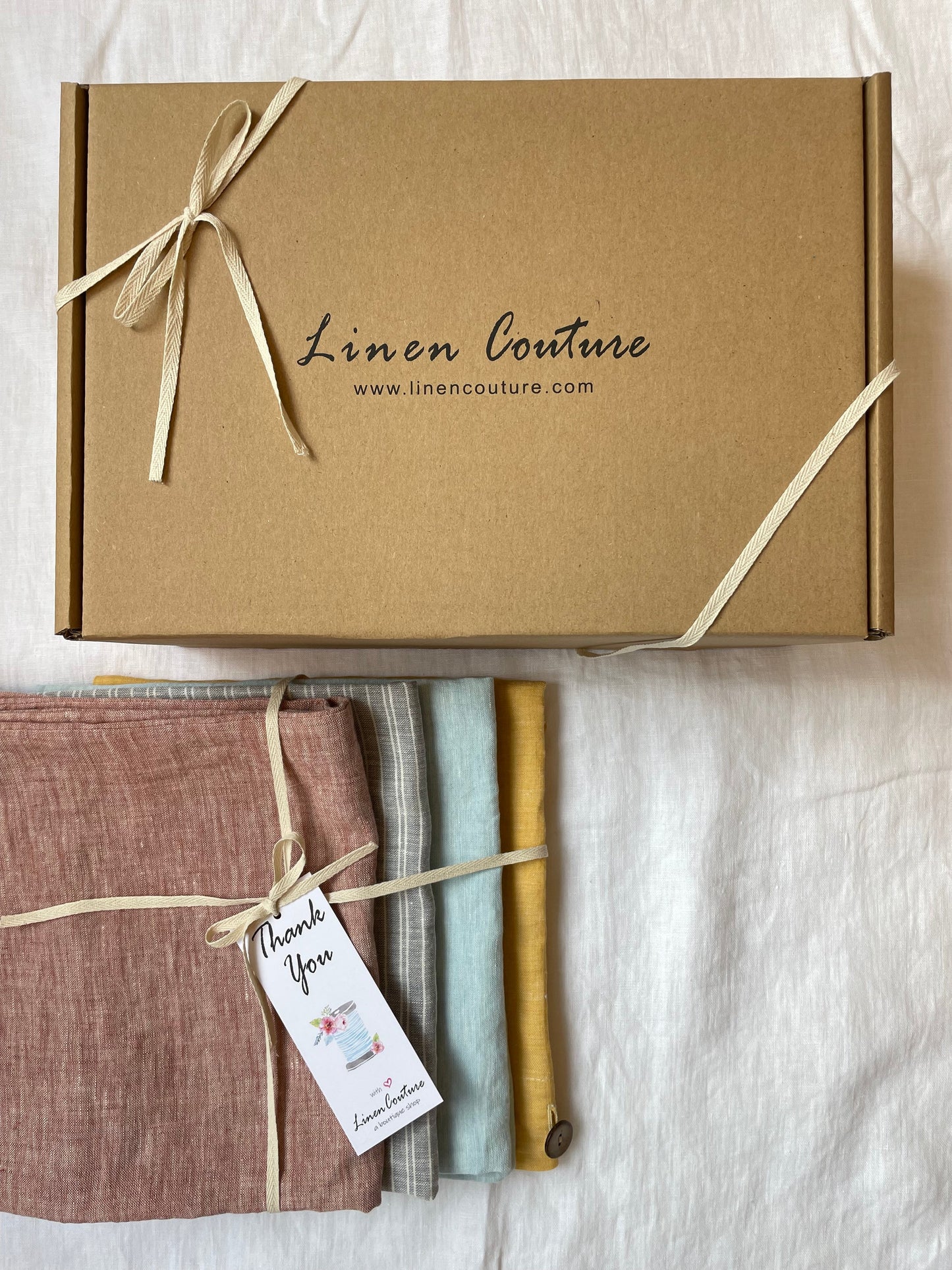 Gift box from Linen Couture with ribbon, accompanied by four folded linen napkins in muted red, grey, aqua, and mustard yellow, tied together with a thank you tag