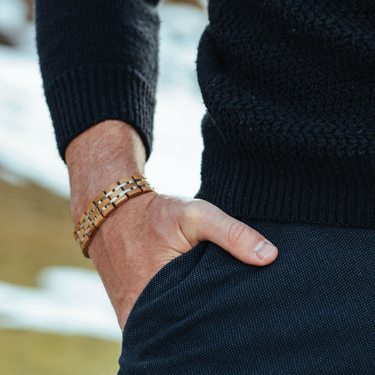 Close-up of a man's wrist wearing a natural wood and stainless steel watch strap, styled with a navy knit sweater and trousers, outdoors in a snowy mountain setting.